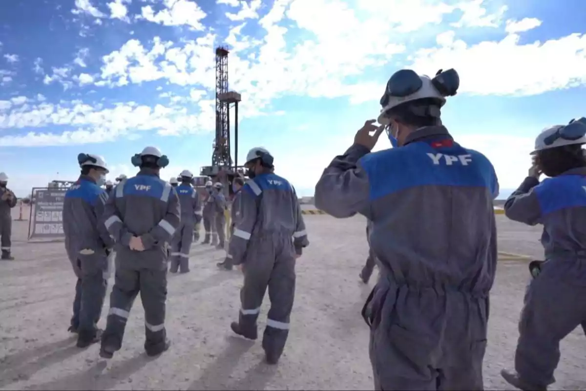 Workers in YPF uniforms walk near an oil platform under a partly cloudy sky.