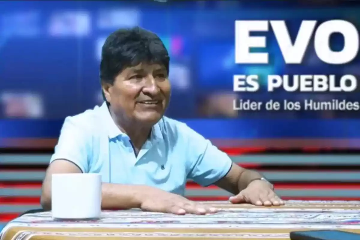Hombre de cabello oscuro y camiseta celeste sentado en una mesa con una taza blanca, al fondo un cartel que dice EVO ES PUEBLO Líder de los Humildes Hombre de cabello oscuro y camiseta celeste sentado en una mesa con una taza blanca, al fondo un cartel que dice EVO ES PUEBLO Líder de los Humildes