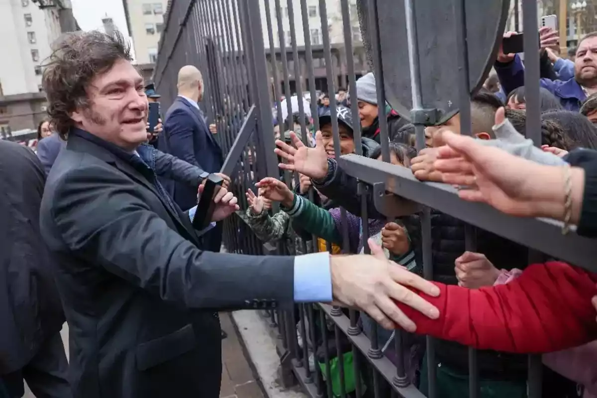 Un hombre sonriente con traje oscuro saluda a un grupo de personas a través de una reja, mientras varias manos se extienden hacia él.