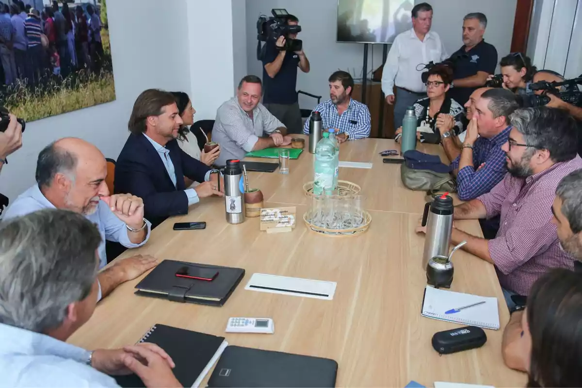 Un grupo de personas se encuentra reunido alrededor de una mesa en una sala de conferencias, algunos de ellos sostienen termos y mates, mientras que varios fotógrafos y camarógrafos documentan el encuentro.