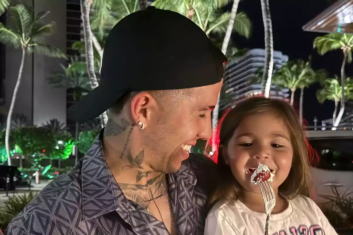 Un hombre con gorra negra y tatuajes sonríe mientras sostiene a una niña que come un postre con un tenedor, con palmeras iluminadas de fondo. Un hombre con gorra negra y tatuajes sonríe mientras sostiene a una niña que come un postre con un tenedor, con palmeras iluminadas de fondo.
