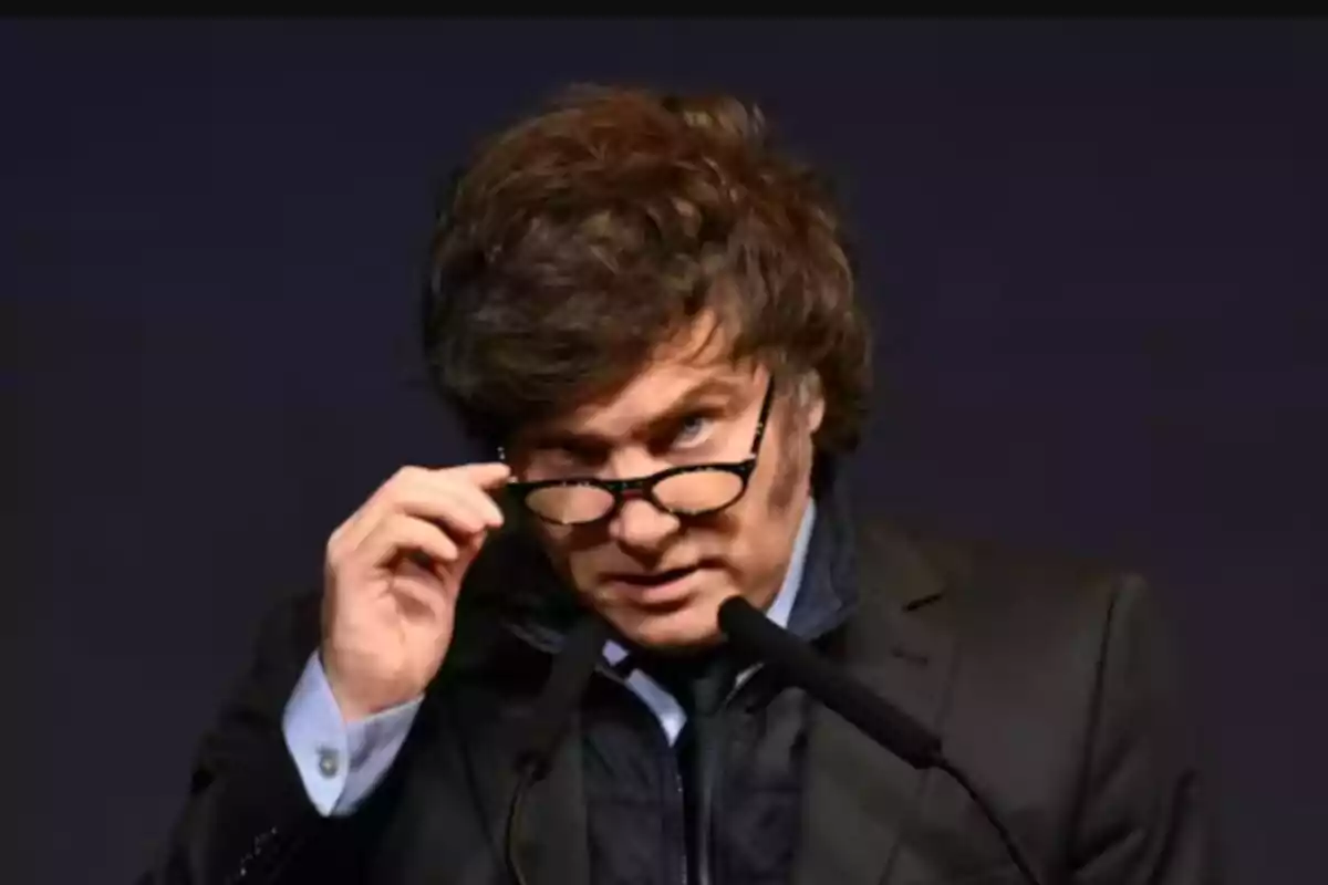 Un hombre con gafas y cabello rizado mira hacia adelante mientras sostiene un micrófono.