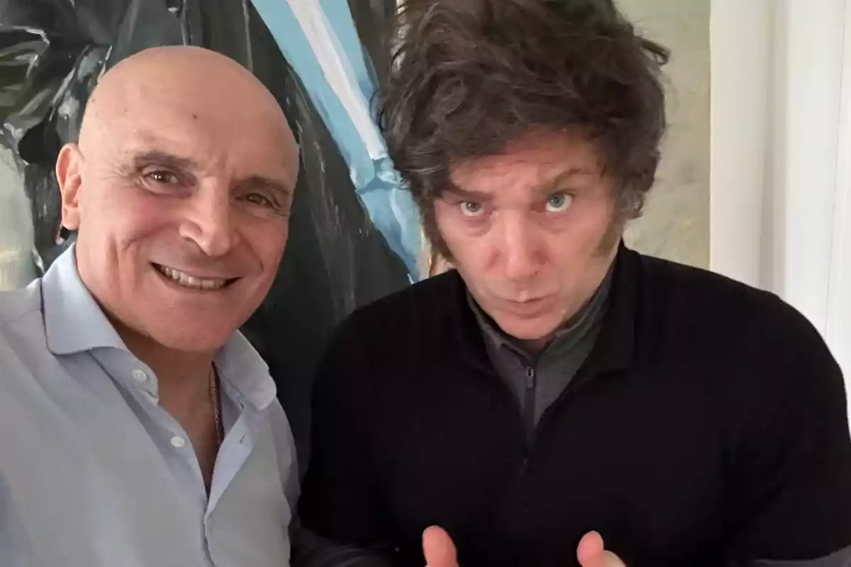 Dos hombres posan juntos para una foto, uno sonríe mientras el otro hace un gesto serio con las manos levantadas, ambos están en un ambiente interior con una pintura de fondo. Dos hombres posan juntos para una foto, uno sonríe mientras el otro hace un gesto serio con las manos levantadas, ambos están en un ambiente interior con una pintura de fondo.