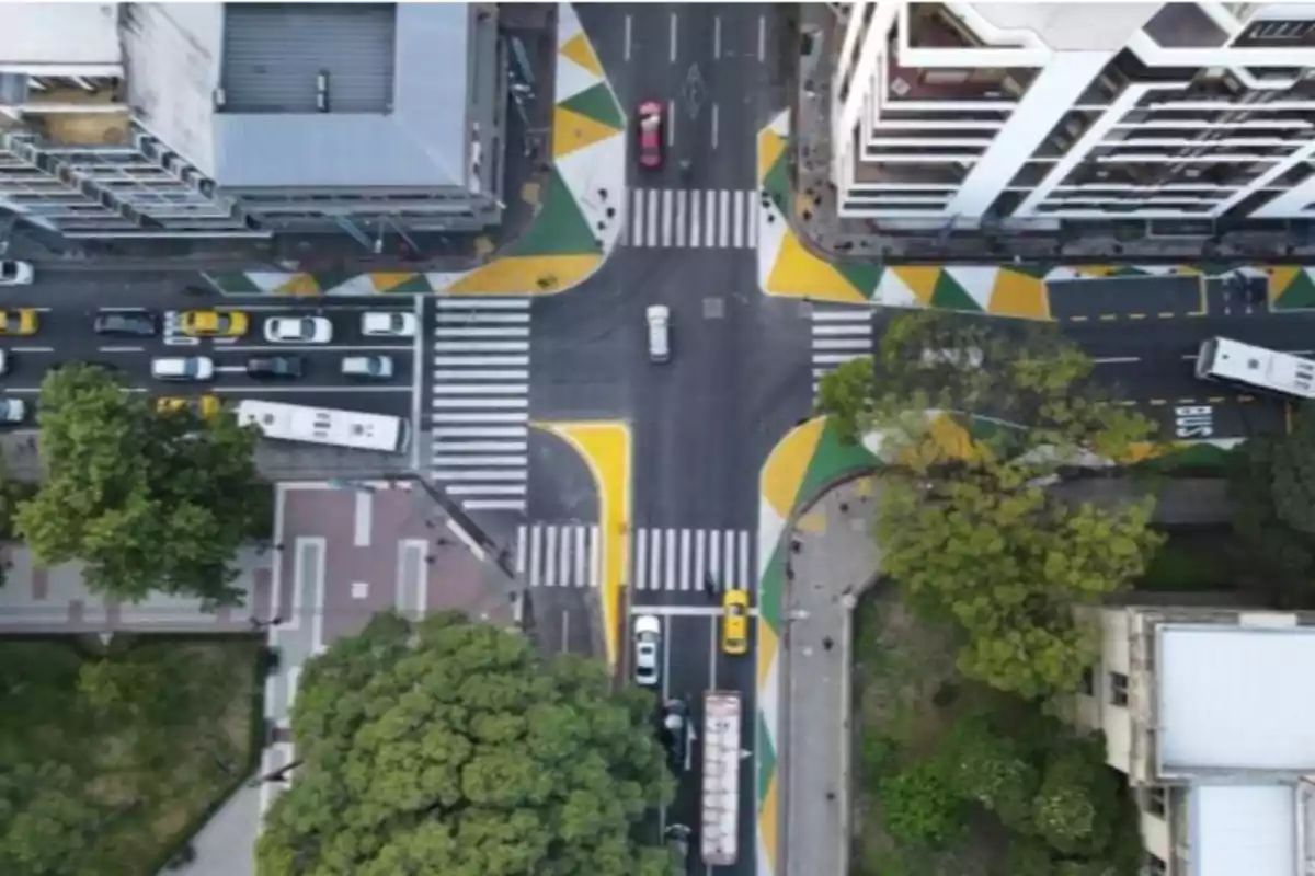 Vista aérea de un cruce de calles con pasos peatonales y esquinas pintadas de amarillo y verde rodeado de edificios, árboles y vehículos circulando