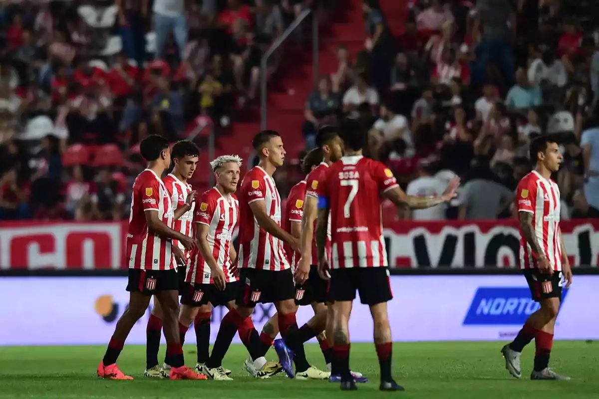 Jugadores de fútbol con camisetas a rayas rojas y blancas en el campo durante un partido, con público en las gradas al fondo.