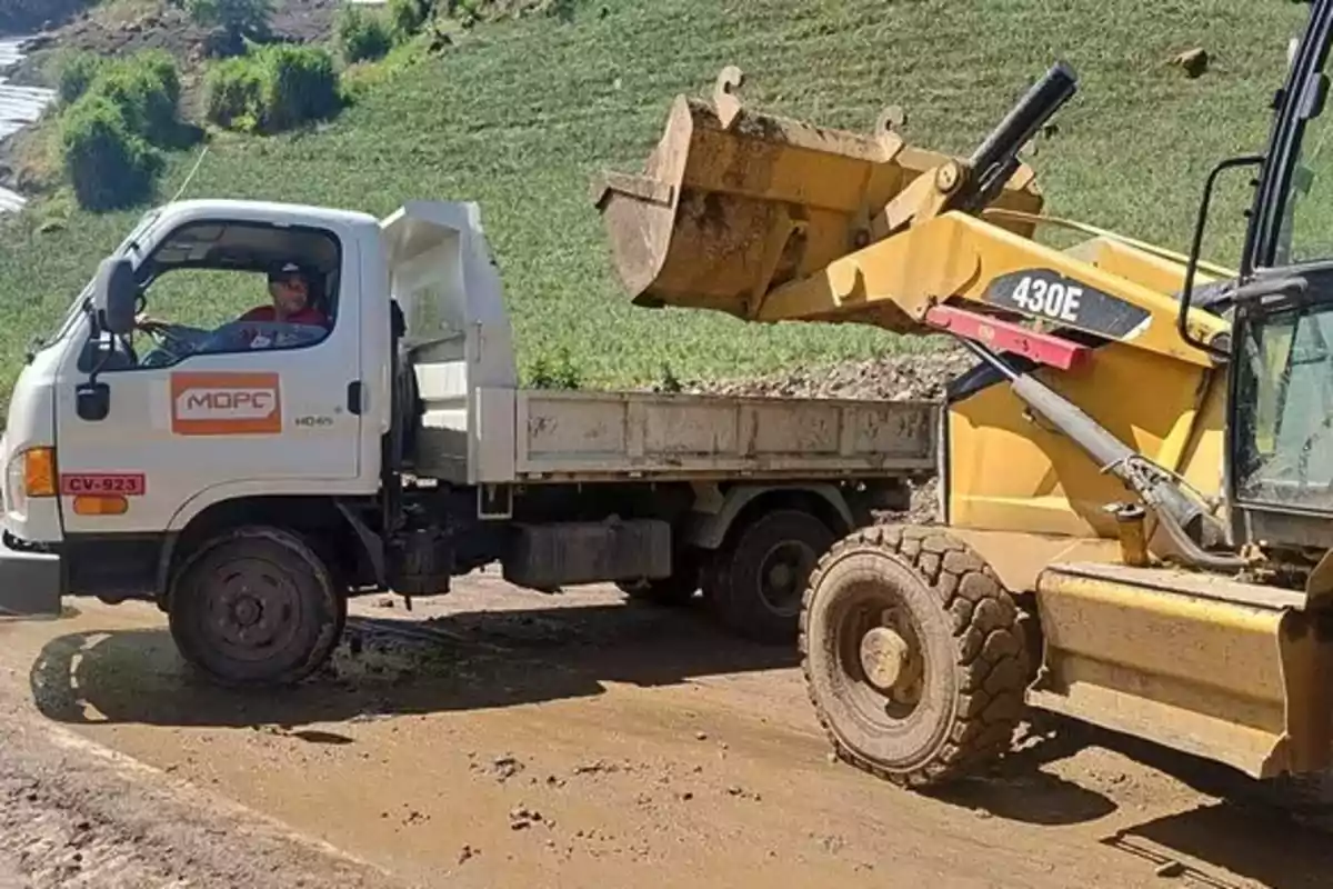 Un camión y una retroexcavadora están trabajando en un camino de tierra, con un paisaje verde al fondo.