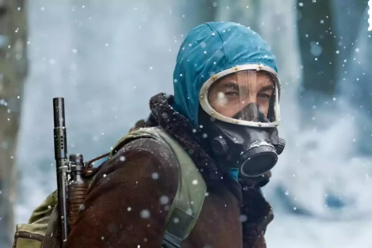 A man with a gas mask and a blue hood in a snowy environment, carrying a backpack and a weapon.
