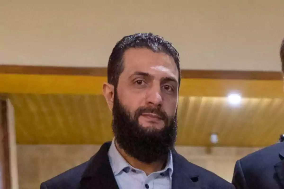 Un hombre con barba y cabello oscuro viste una chaqueta negra y camisa clara en un entorno interior con techo de madera.