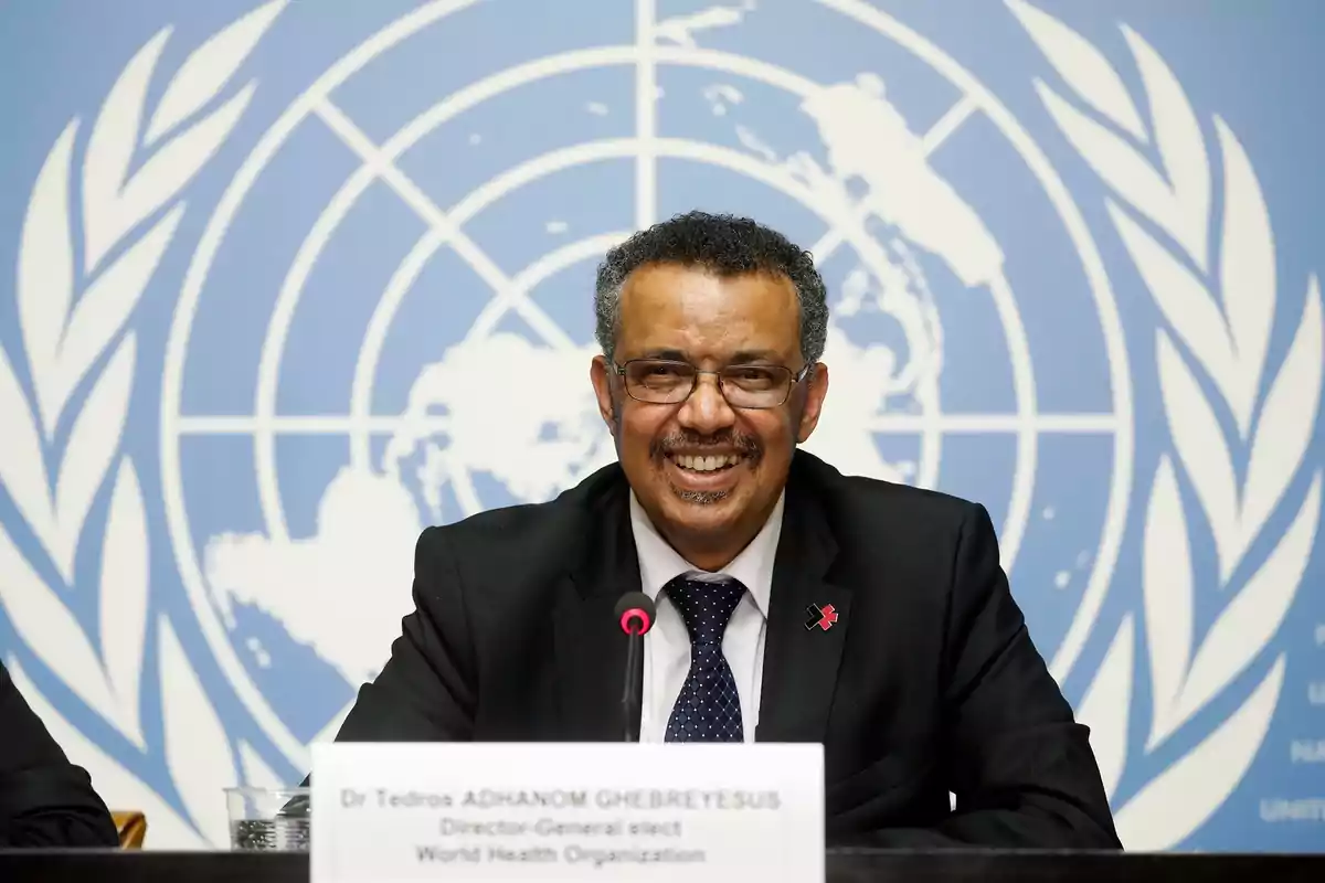 Un hombre sonriente con gafas y traje oscuro sentado frente a un micrófono con un fondo azul que tiene el logotipo de la ONU.