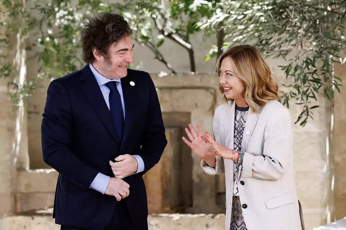 Dos personas conversando y sonriendo en un entorno al aire libre con árboles y vegetación de fondo.