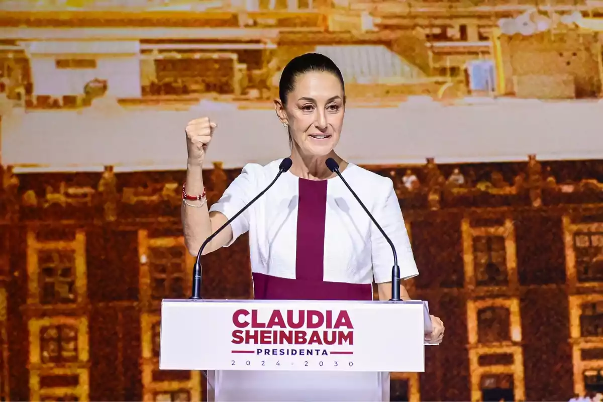 Una mujer en un podio con un fondo urbano, levantando el puño mientras da un discurso.