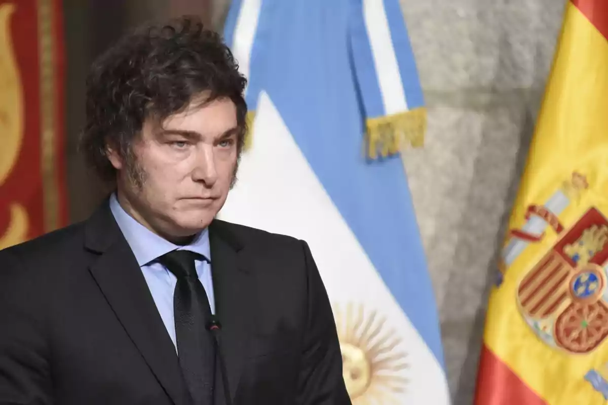 Un hombre con traje oscuro y corbata negra está de pie frente a las banderas de Argentina y España.