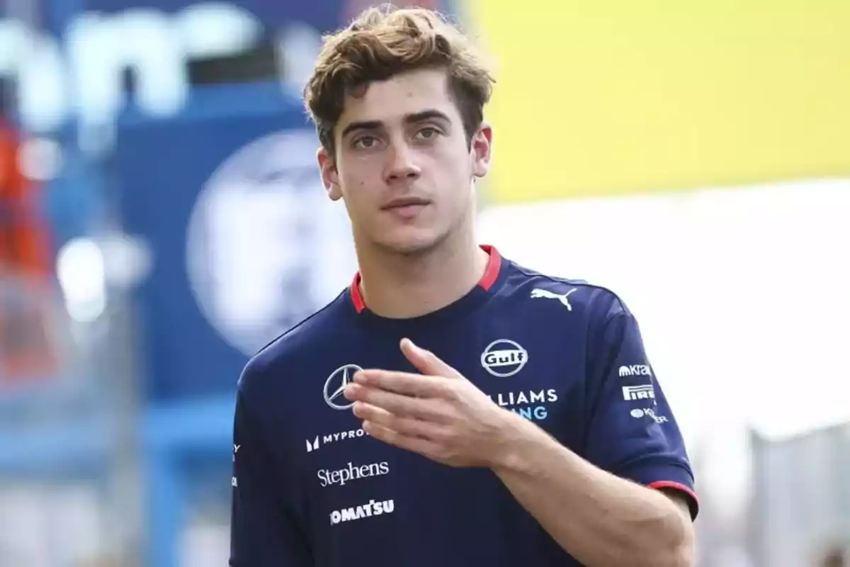 Un joven con uniforme de carreras azul oscuro con logotipos de patrocinadores en un entorno al aire libre.