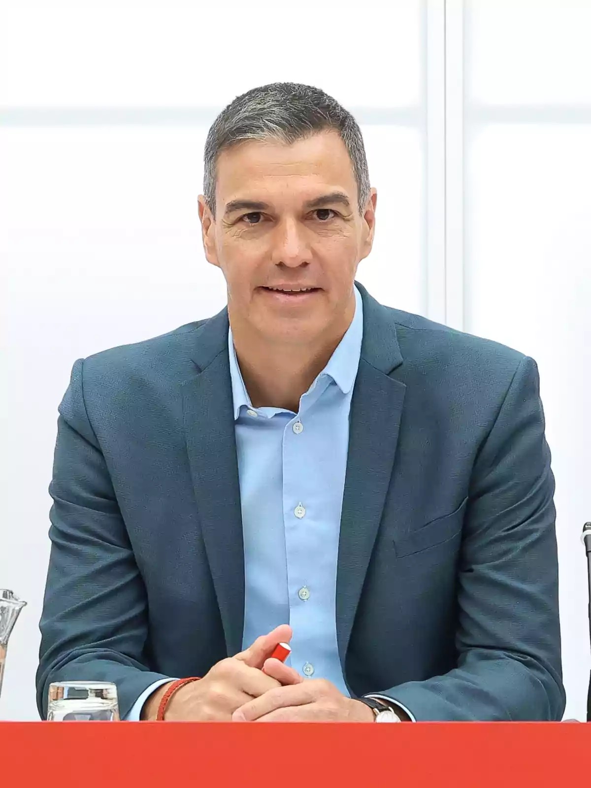 Hombre con traje azul claro y camisa azul sentado frente a una mesa roja.