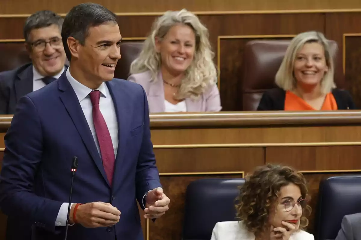 Un hombre de traje azul y corbata roja habla en un parlamento mientras varias personas sonríen en el fondo.