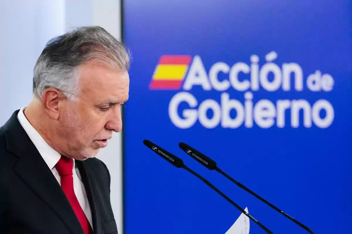 Un hombre de traje y corbata roja habla frente a un micrófono con un cartel azul de fondo que dice 