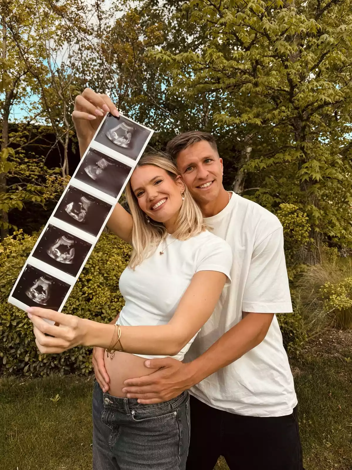 A smiling couple in a garden, the woman holding a series of ultrasounds while the man hugs her by the waist. A smiling couple in a garden, the woman holding a series of ultrasounds while the man hugs her by the waist.