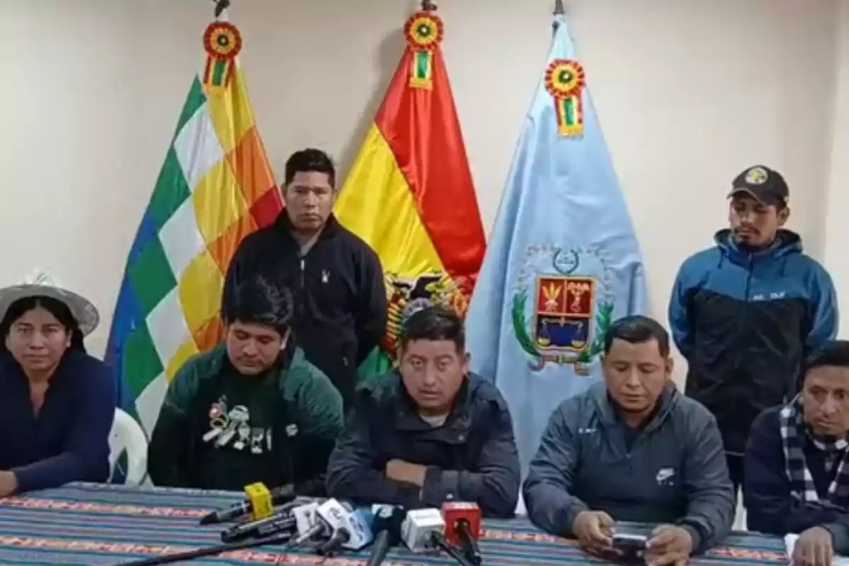 Un grupo de personas sentadas y de pie frente a tres banderas bolivianas durante una conferencia de prensa, con micrófonos sobre la mesa.