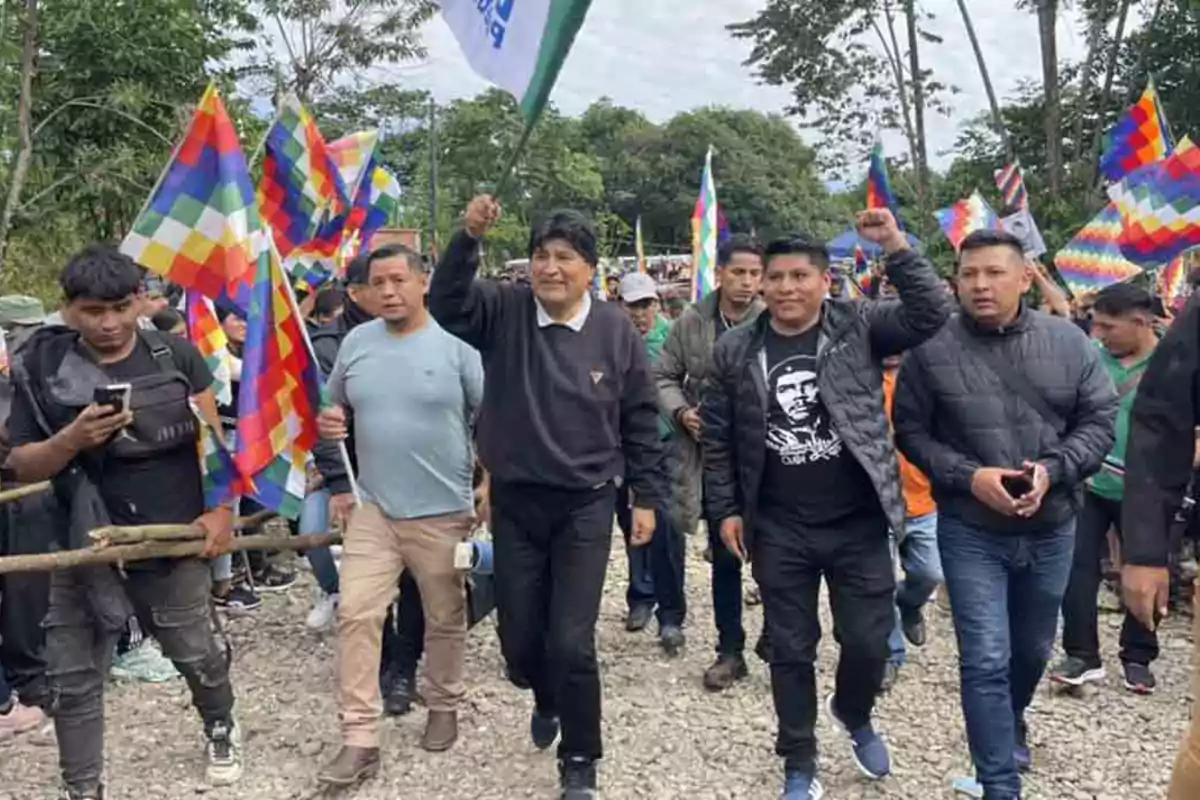 Un grupo de personas marcha por un camino de tierra sosteniendo banderas multicolores y una bandera blanca rodeados de árboles y vegetación