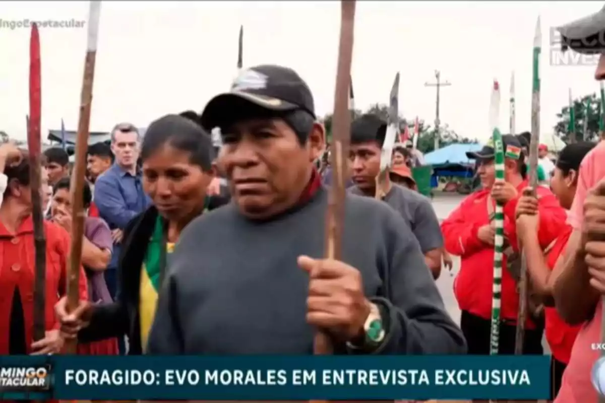 Un grupo de personas indígenas sostiene palos mientras participan en una manifestación, con un ambiente de protesta y varias personas observando al fondo