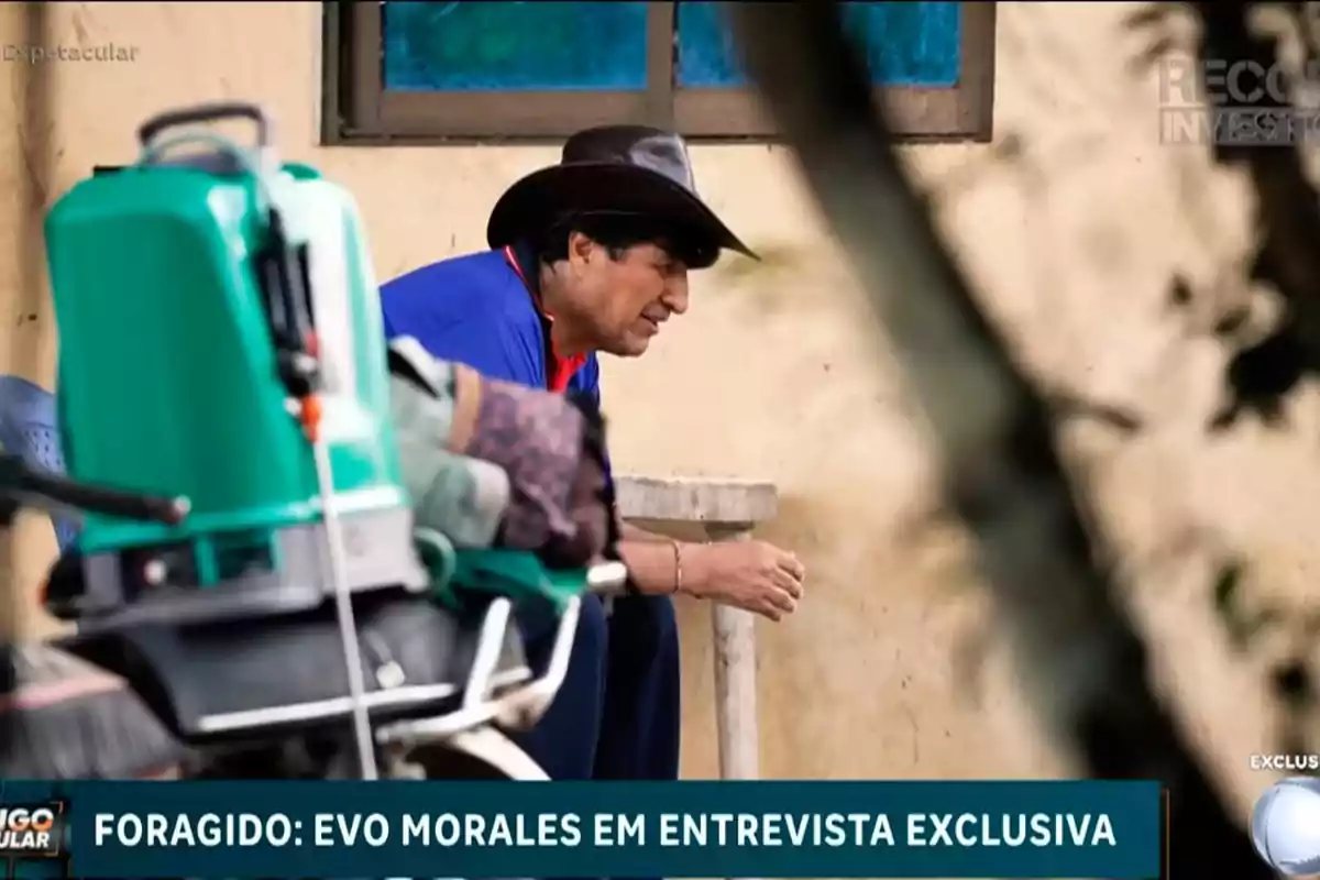 Man with a black hat and blue jersey sitting next to a concrete table in an outdoor setting, with a green machine in the foreground and a sign at the bottom mentioning an exclusive interview.