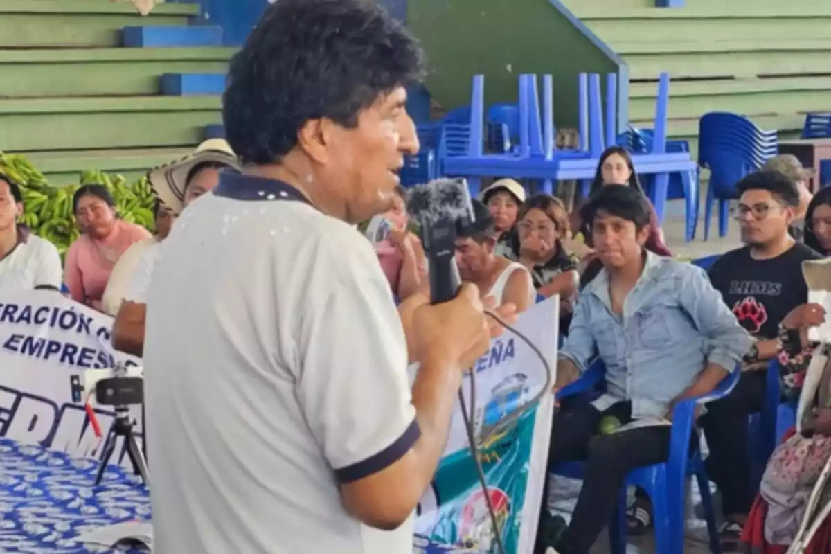 Un hombre hablando con un micrófono frente a un grupo de personas sentadas en sillas azules en un espacio cubierto.