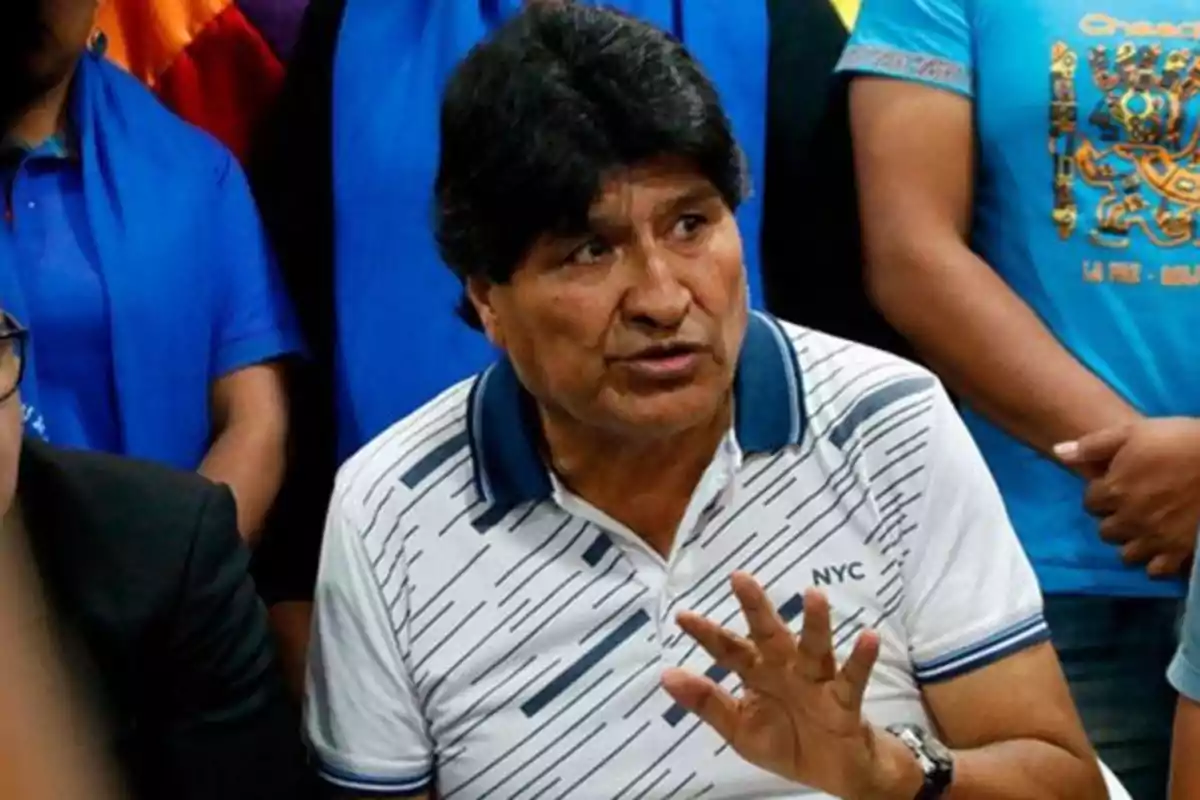 Un hombre de cabello oscuro y camisa blanca con rayas habla gesticulando con la mano, rodeado de personas con camisetas azules.