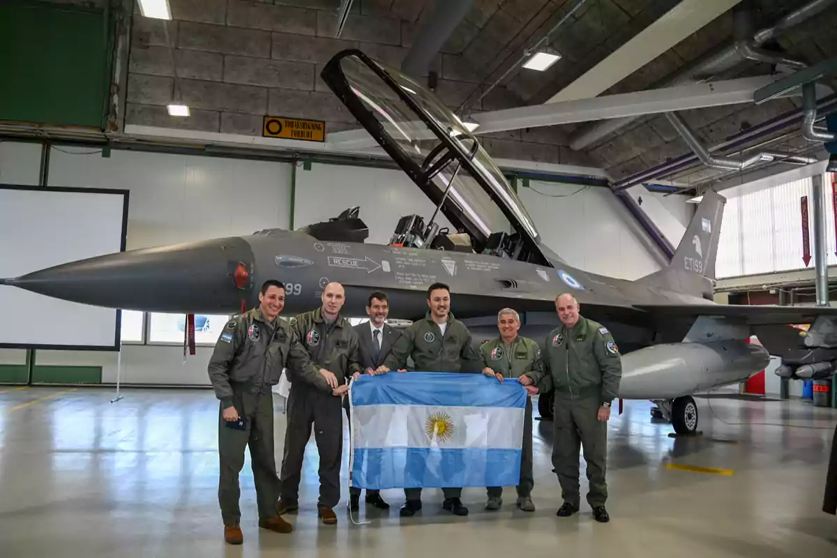 Seis hombres posan sonrientes frente a un avión de combate en un hangar mientras sostienen una bandera de Argentina