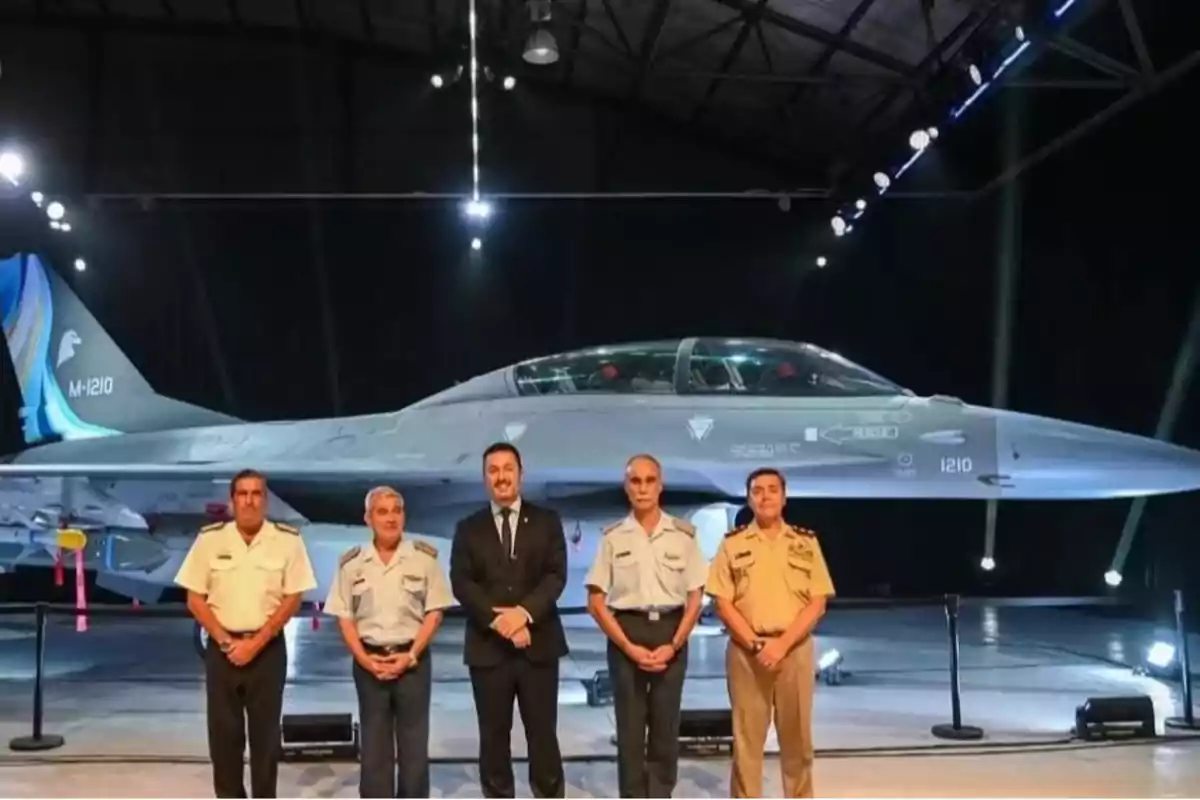 Five men pose in front of a fighter jet at an indoor exhibition with lights focused on the aircraft.