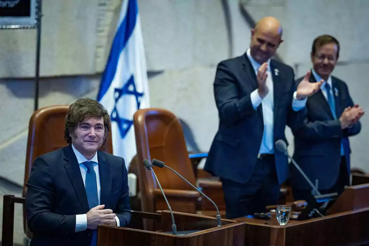 Un hombre de traje está de pie en un podio mientras dos personas lo aplauden y una bandera de Israel se ve al fondo