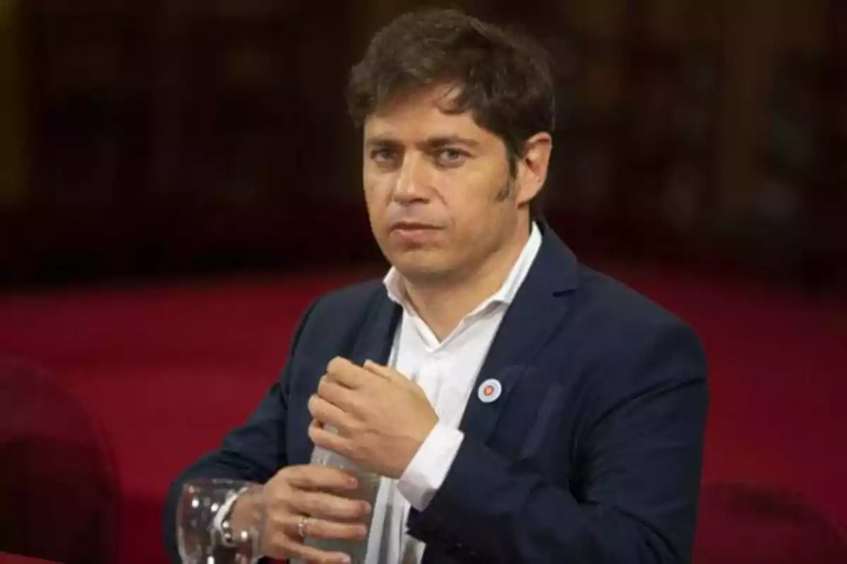 Hombre con traje oscuro y camisa blanca sentado en una mesa con fondo borroso rojo.