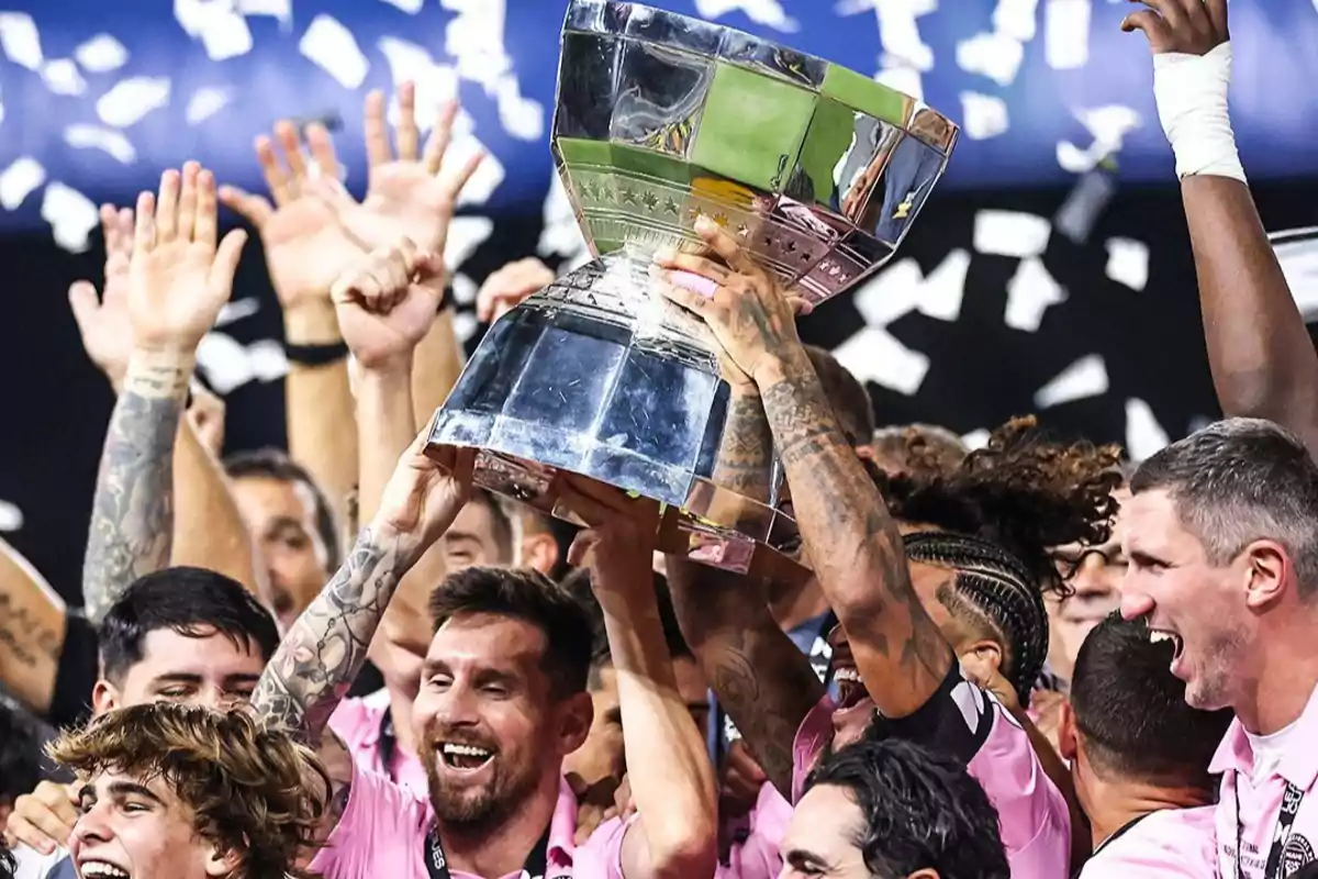 Jugadores de fútbol con uniforme rosa celebran levantando un trofeo plateado mientras caen papeles de confeti blancos Jugadores de fútbol con uniforme rosa celebran levantando un trofeo plateado mientras caen papeles de confeti blancos