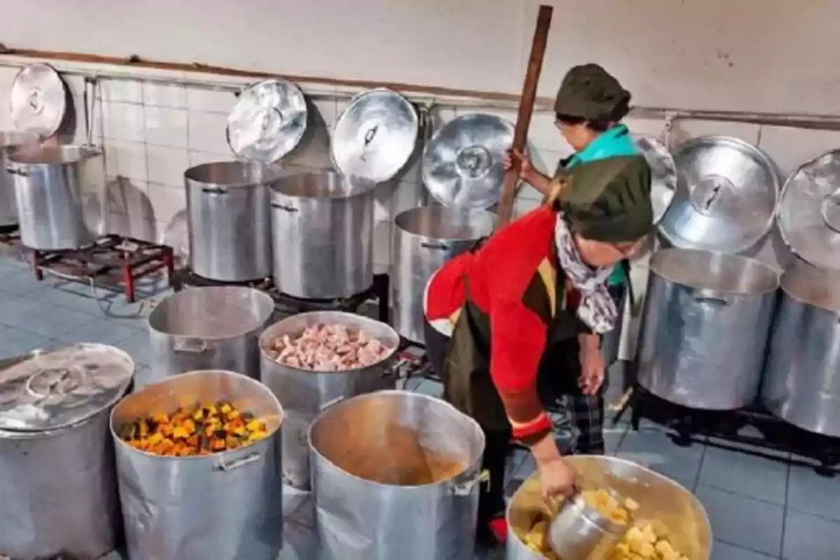 Dos personas cocinando en una cocina industrial con grandes ollas de metal llenas de ingredientes.