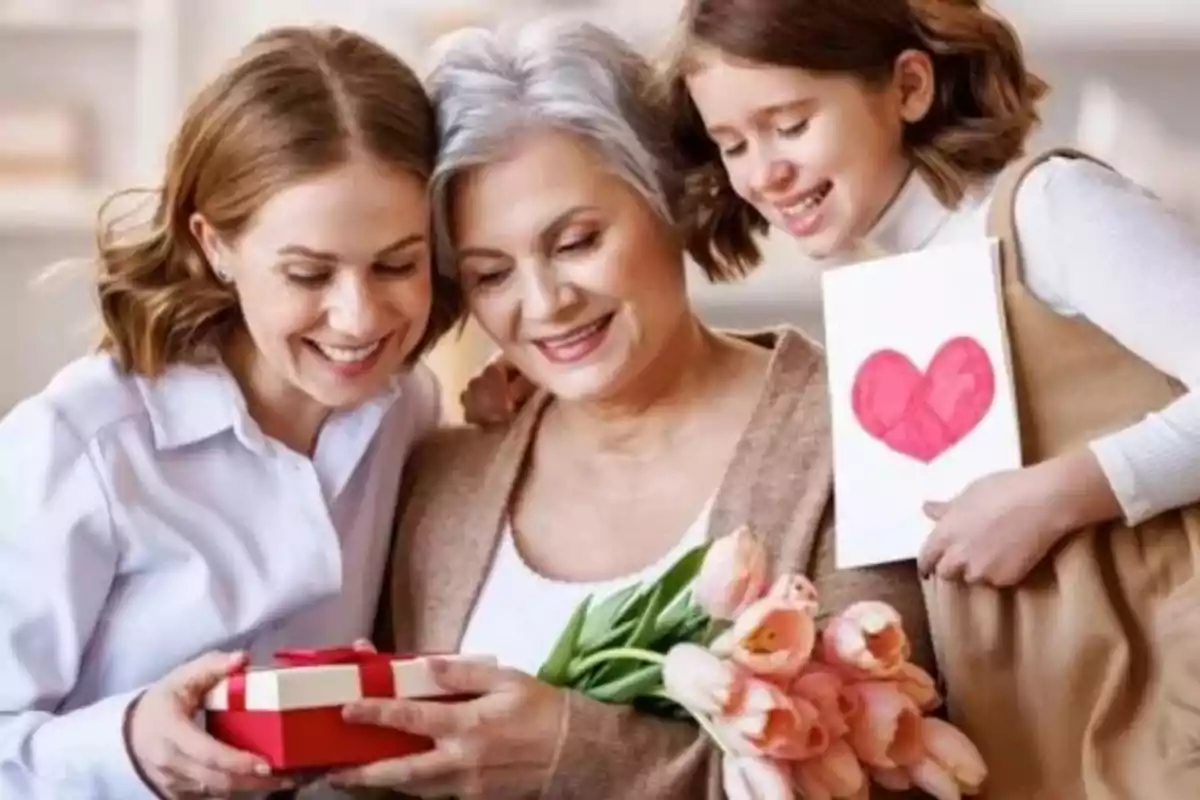 Tres generaciones de mujeres sonríen mientras sostienen un regalo, flores y una tarjeta con un corazón.