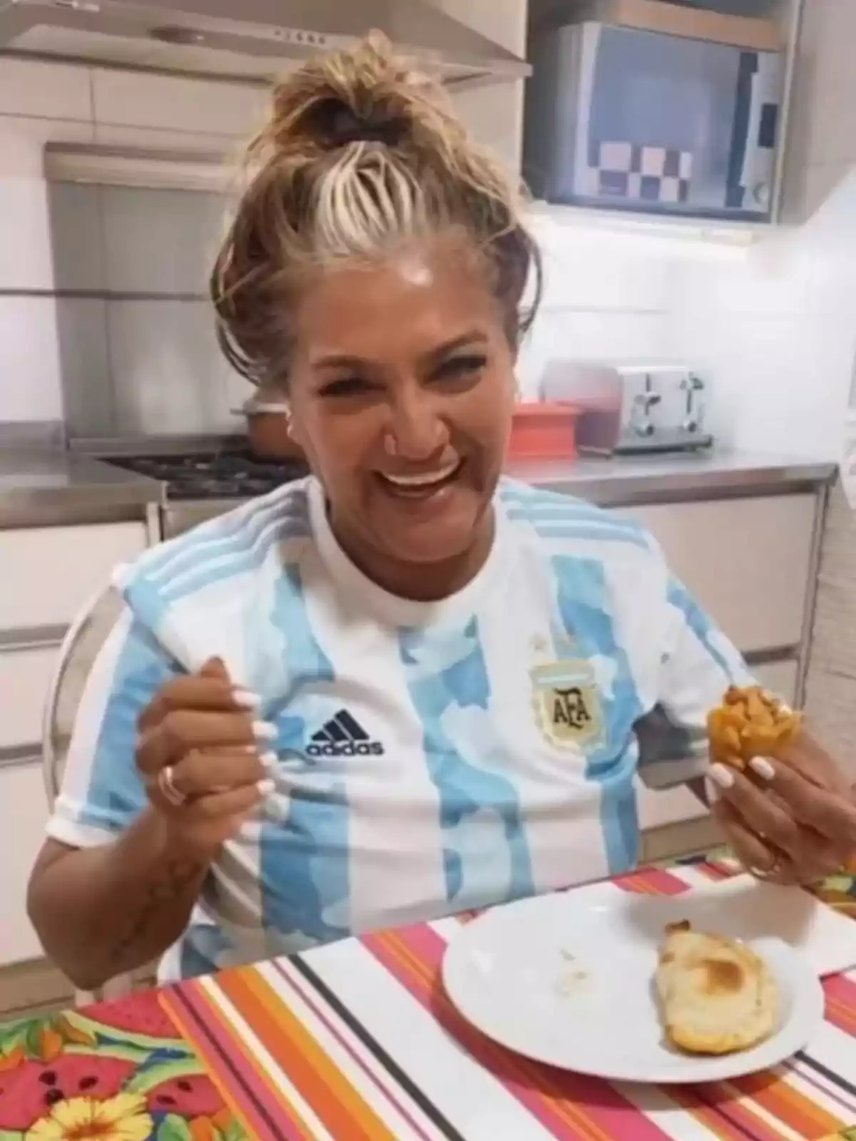 A smiling person wearing an Argentina soccer jersey is sitting in a kitchen, holding an empanada in one hand and a fork in the other, in front of a plate on a colorful tablecloth. A smiling person wearing an Argentina soccer jersey is sitting in a kitchen, holding an empanada in one hand and a fork in the other, in front of a plate on a colorful tablecloth.