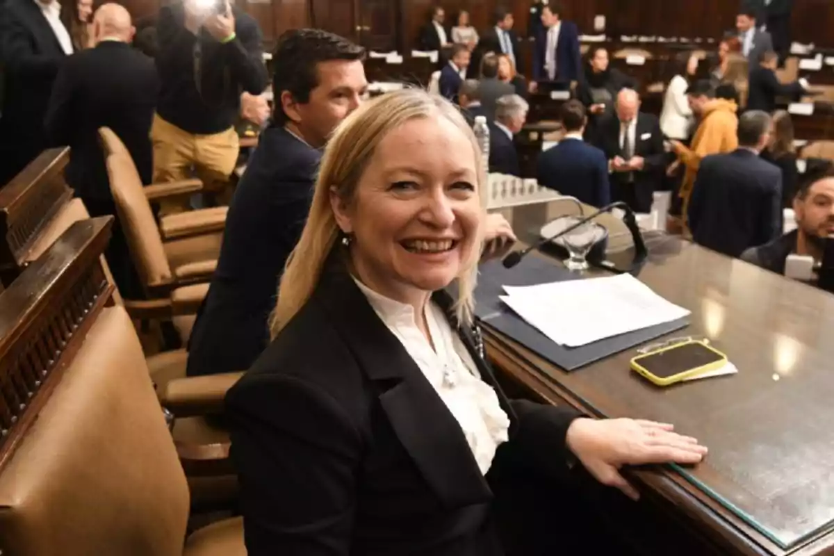 A smiling woman sitting at a desk in a formal setting with several people in the background. A smiling woman sitting at a desk in a formal setting with several people in the background.