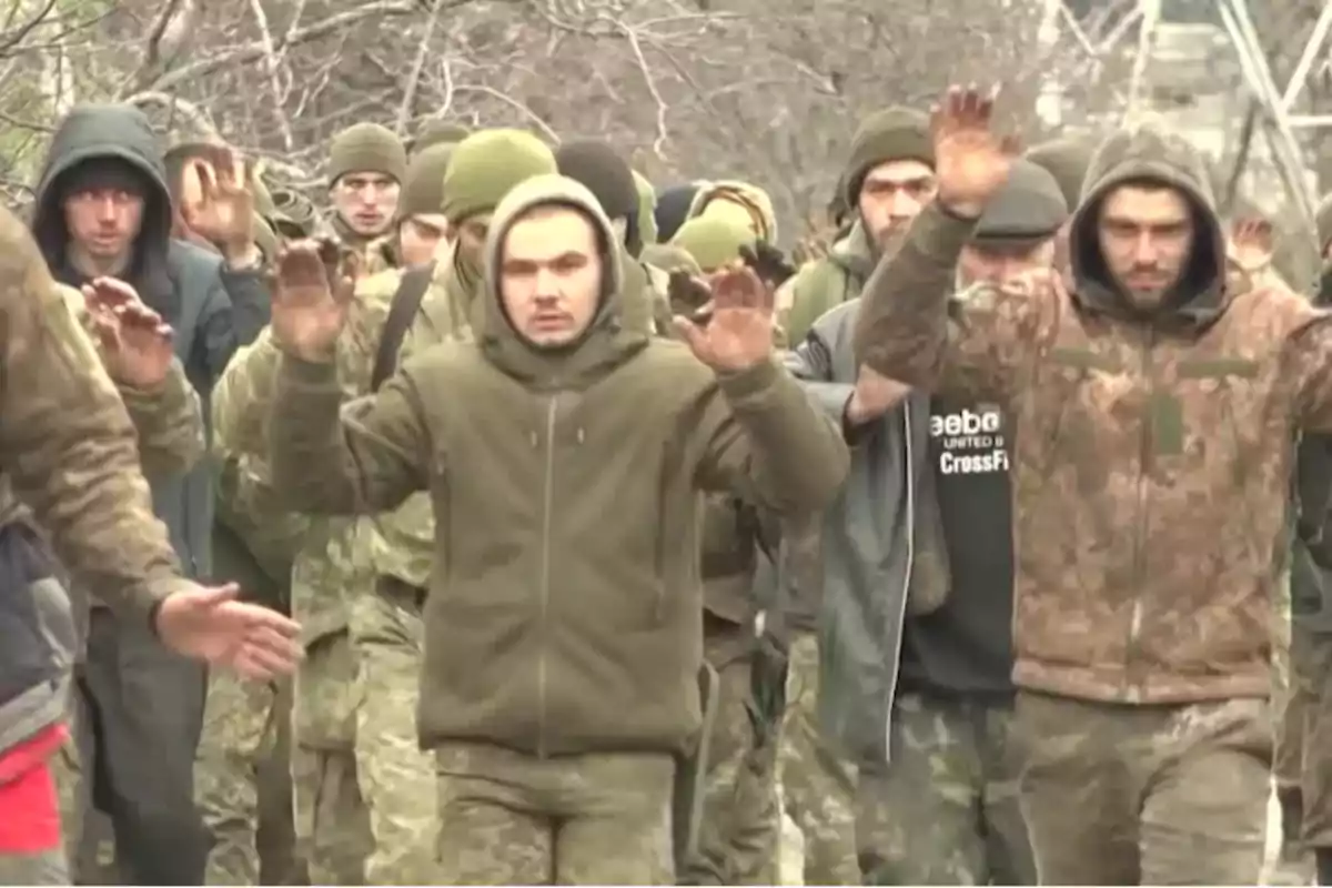 A group of people in camouflage clothing and coats walks with their hands raised in an outdoor setting. A group of people in camouflage clothing and coats walks with their hands raised in an outdoor setting.
