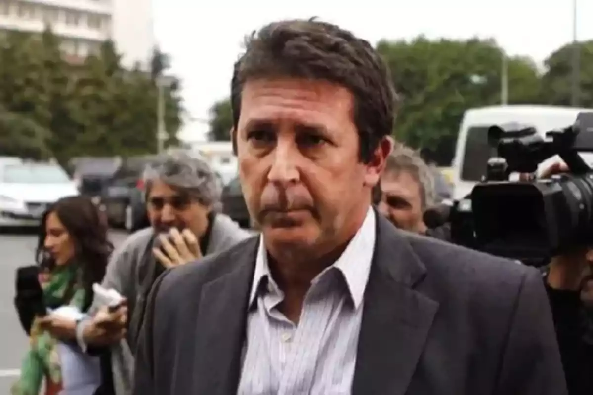 Short dark-haired man with a serious expression wearing a jacket and light-colored jersey, surrounded by people and cameras in an urban setting.