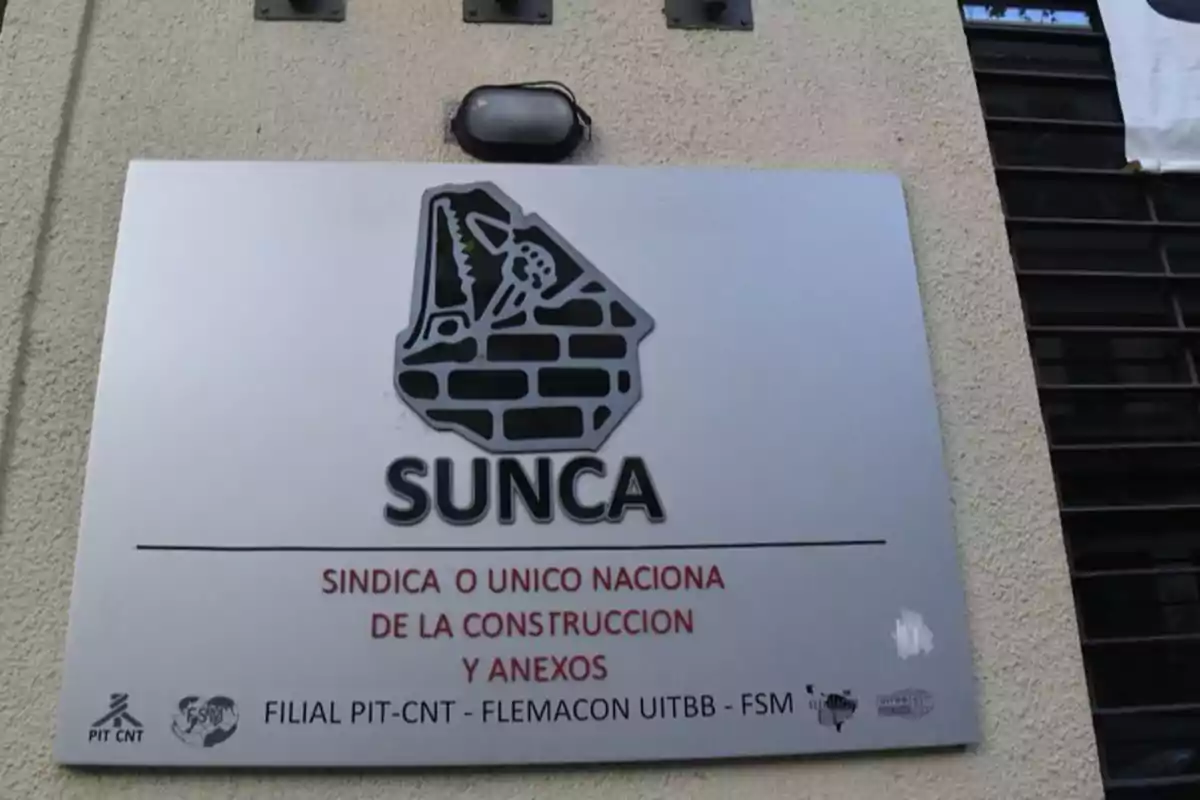Cartel metálico en la pared de un edificio con el logo de un muro de ladrillos y herramientas, el texto SUNCA y la leyenda Sindicato Único Nacional de la Construcción y Anexos junto a varios logotipos pequeños en la parte inferior Cartel metálico en la pared de un edificio con el logo de un muro de ladrillos y herramientas, el texto SUNCA y la leyenda Sindicato Único Nacional de la Construcción y Anexos junto a varios logotipos pequeños en la parte inferior