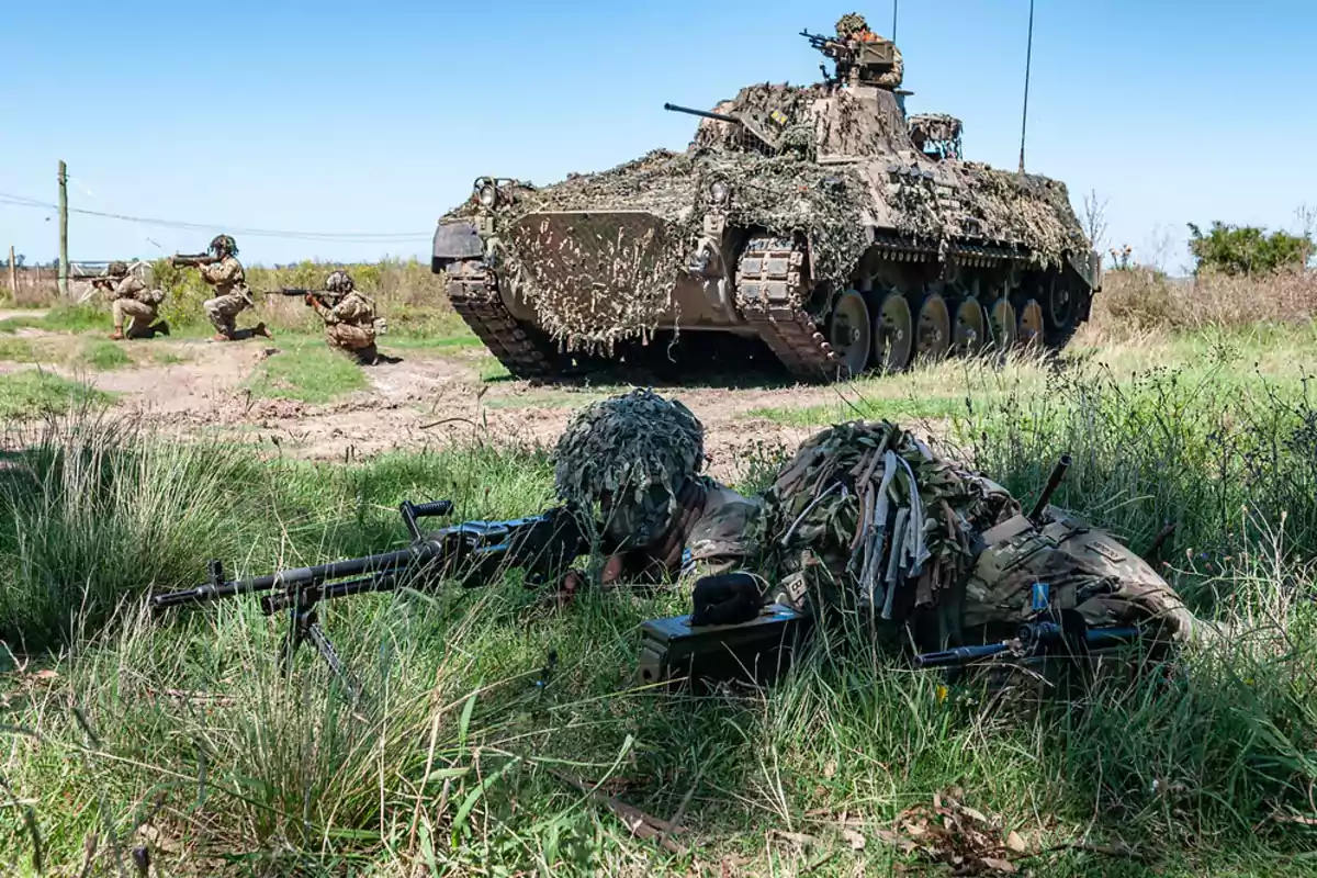 Soldados camuflados en un ejercicio militar con un vehículo blindado en un campo abierto.