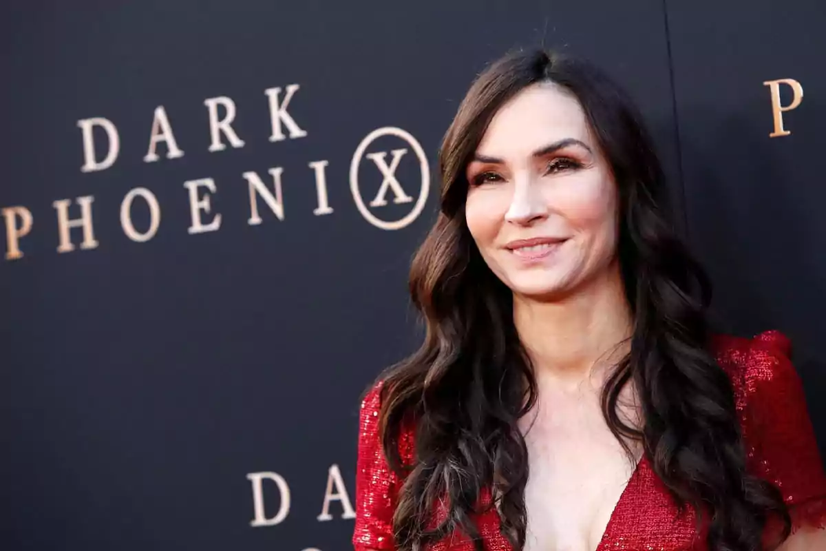 Woman with long dark hair in a red dress smiles in front of a promotional backdrop for the movie Dark Phoenix Woman with long dark hair in a red dress smiles in front of a promotional backdrop for the movie Dark Phoenix