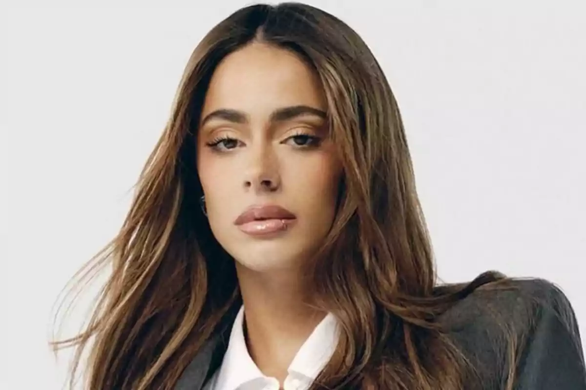 Una persona con cabello largo y castaño vistiendo una chaqueta gris y camisa blanca.
