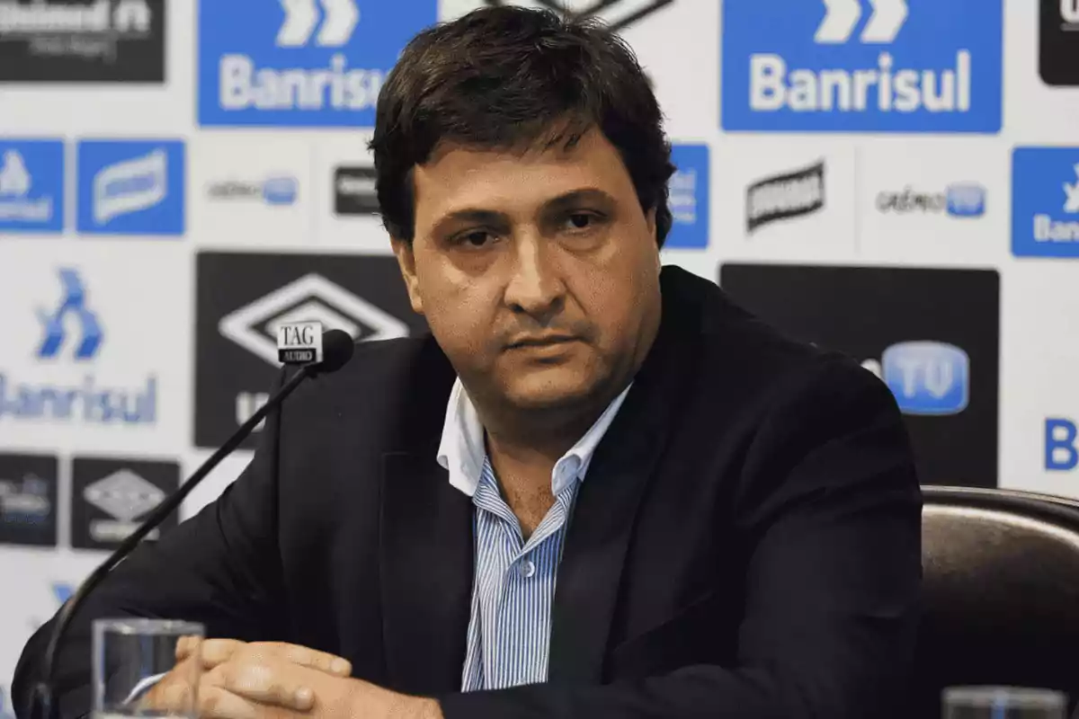 A man at a press conference with sponsor logos in the background. A man at a press conference with sponsor logos in the background.