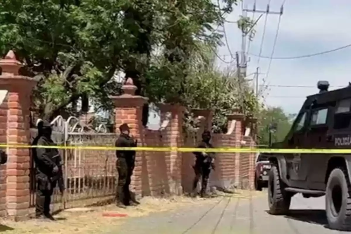 Heavily armed police officers guard a house behind yellow security tape next to an armored patrol vehicle on a tree-lined street.