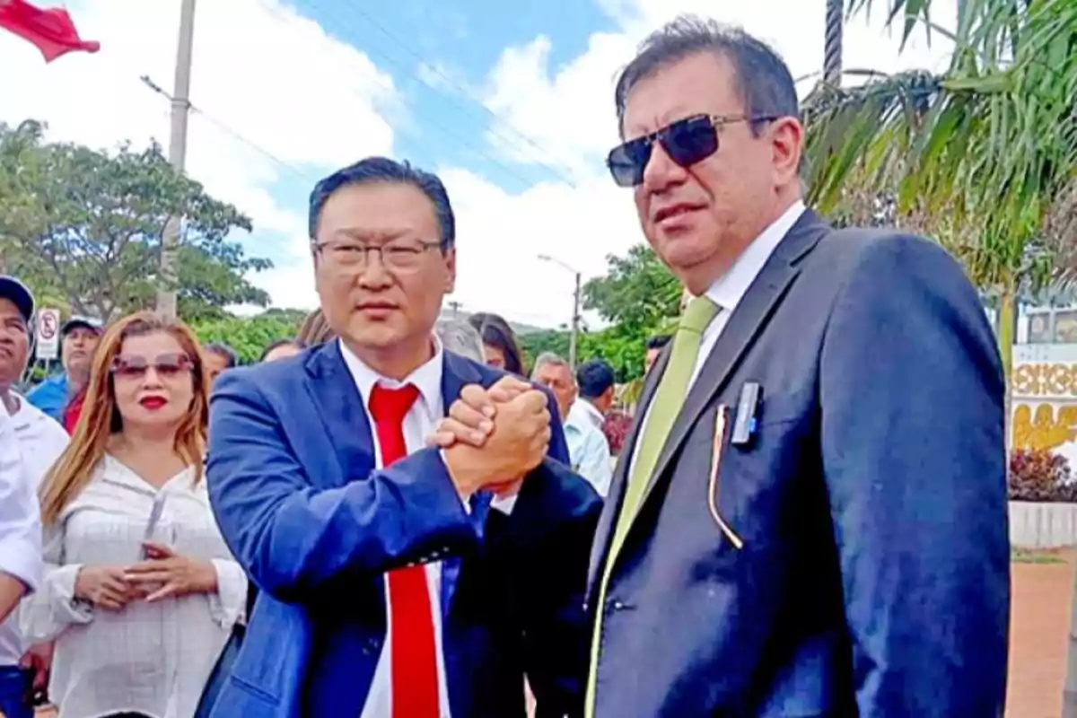 Dos hombres vestidos de traje se dan la mano en un evento al aire libre, rodeados de otras personas.