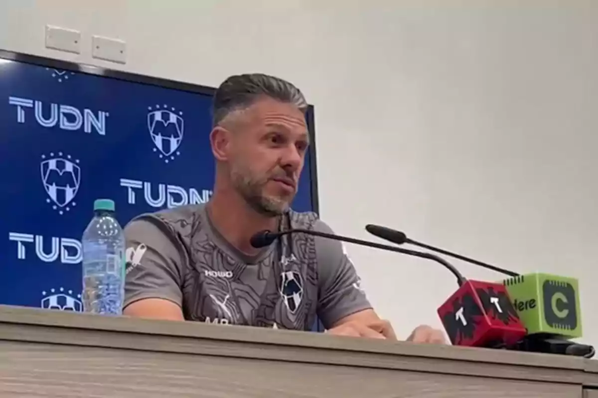 A man in a sports jersey speaks at a press conference, with microphones and a background screen displaying a soccer team's logo. A man in a sports jersey speaks at a press conference, with microphones and a background screen displaying a soccer team's logo.