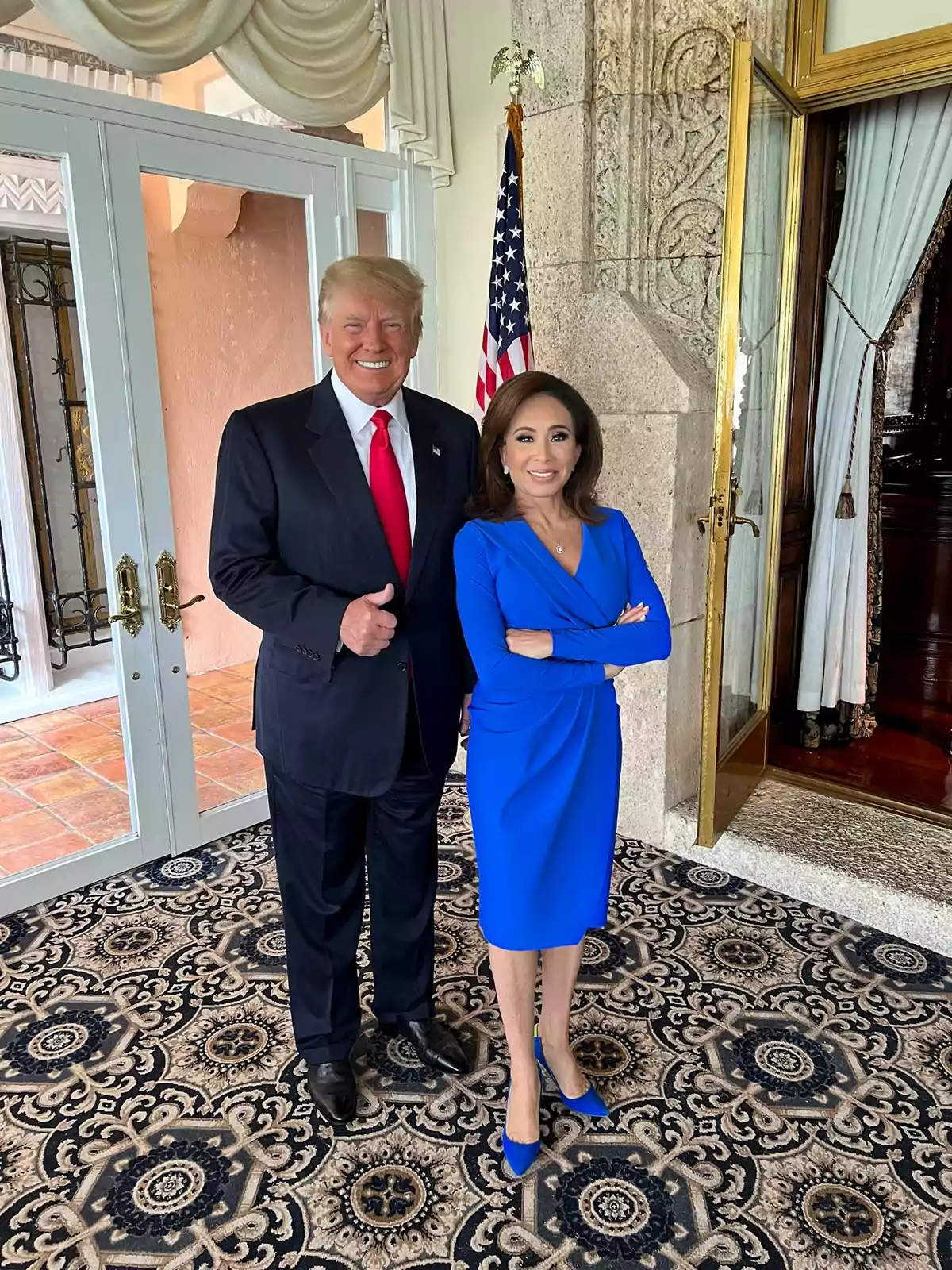 Dos personas posan juntas en un interior elegante con una alfombra decorativa y una bandera de Estados Unidos al fondo. Dos personas posan juntas en un interior elegante con una alfombra decorativa y una bandera de Estados Unidos al fondo.