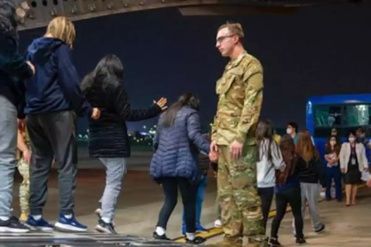 A group of people disembarks from a military plane at an airport at night, guided by a soldier.