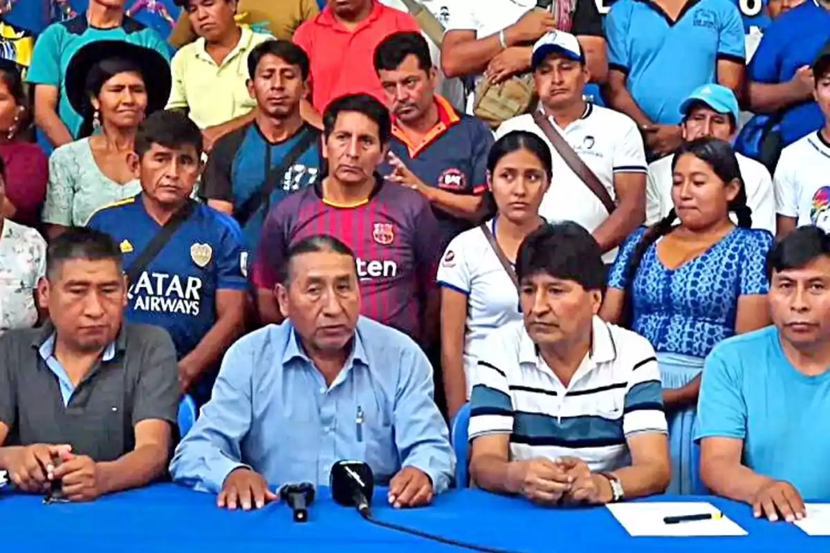 Un grupo de personas está reunido alrededor de una mesa con un micrófono, algunas de ellas están sentadas y otras de pie al fondo.