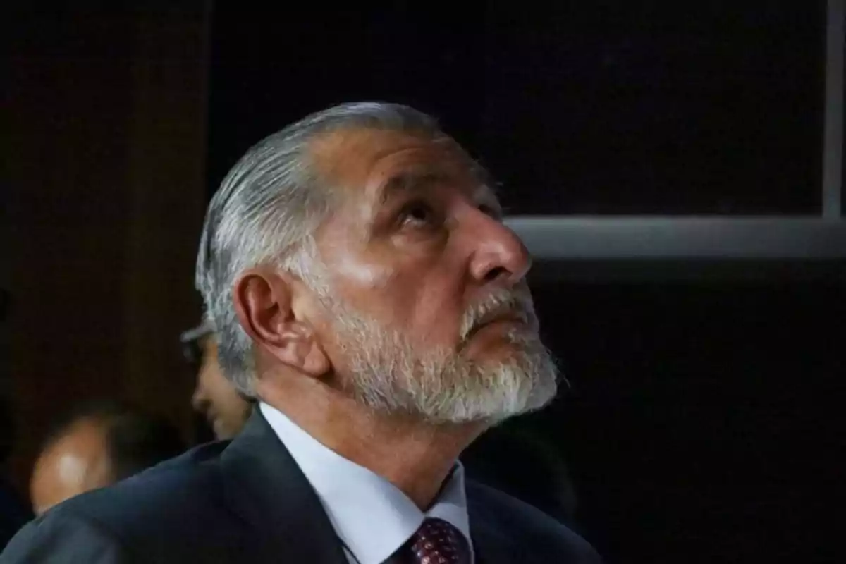 A gray-haired man with a beard looks up in a dark setting.